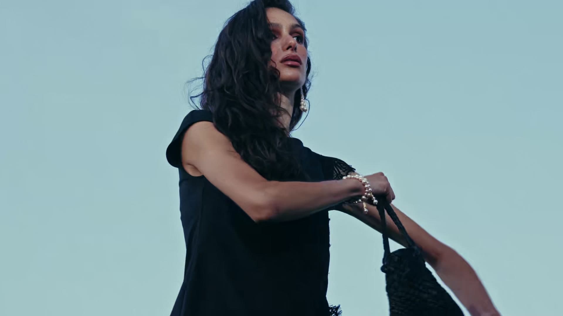 Model in a black dress with a woven bag against a pale sky, on set by the sea